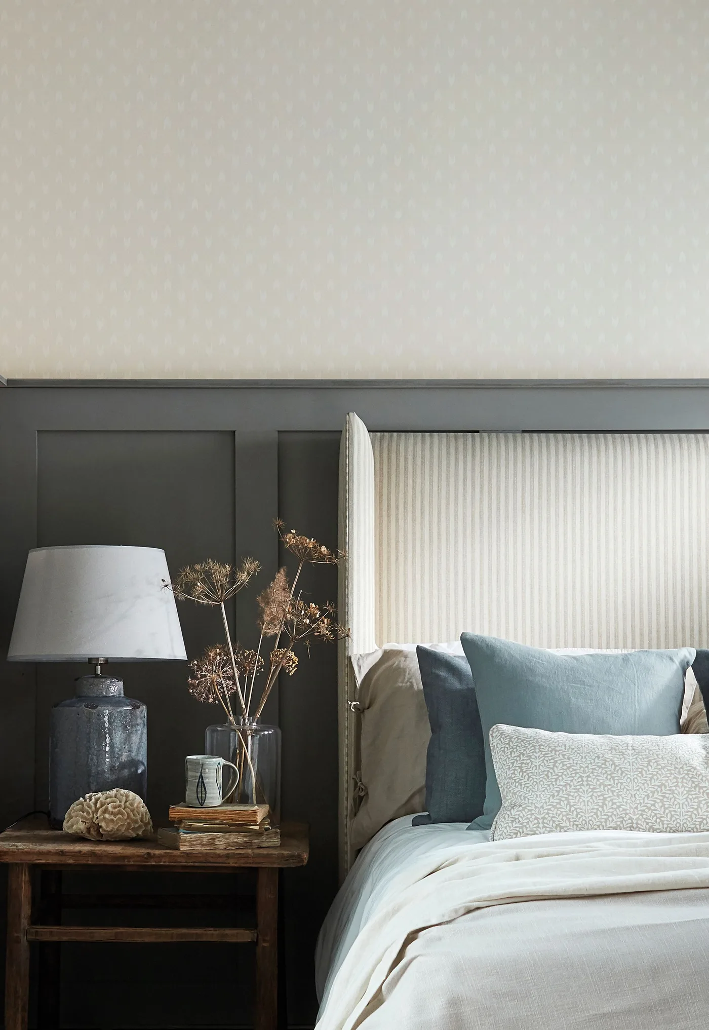 Bedroom detail with dark gray lower wall paneling, pale striped winged headboard, blue cushion and wooden bedside table.