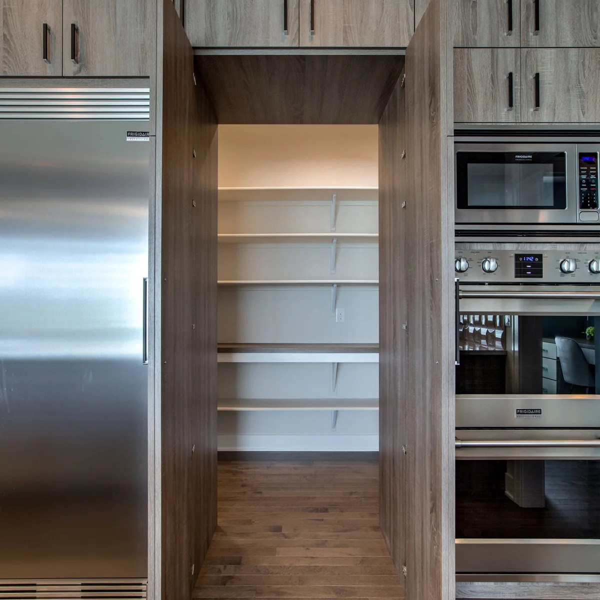 Tall wood-look doors stand open between a refrigerator and ovens to reveal a walk-in pantry with white wall shelves.