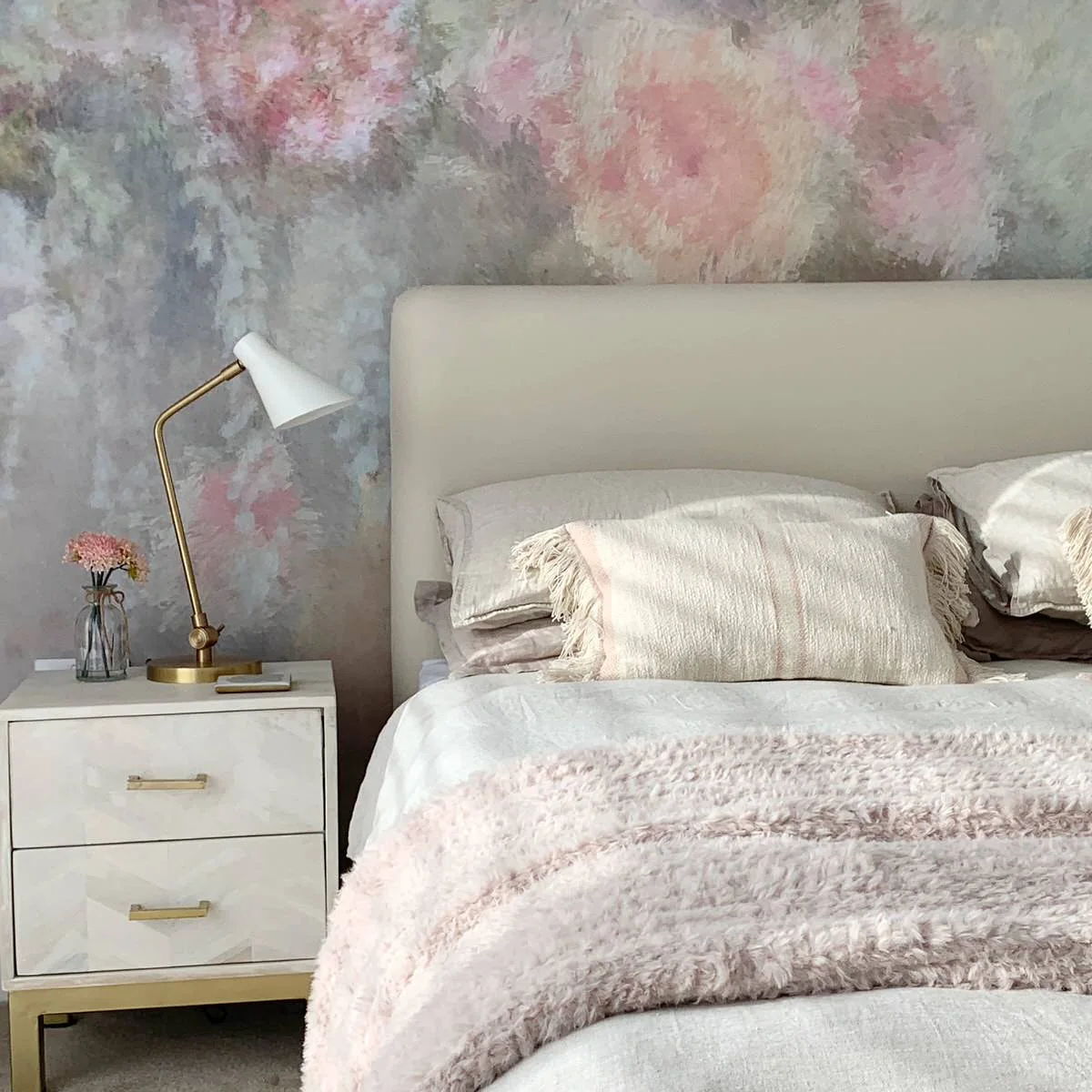 Cream headboard and white bedside drawers against a softly mottled pink-and-blue wall with pale bedding