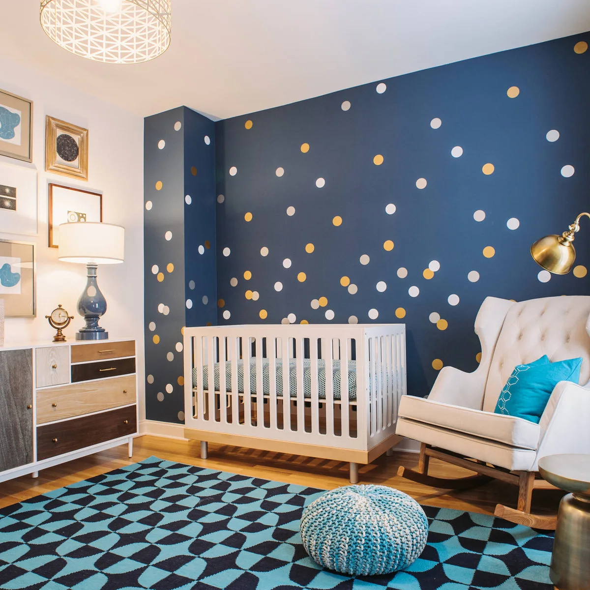 White crib against a dark blue wall with white and gold-colored dots beside a pale wingback chair