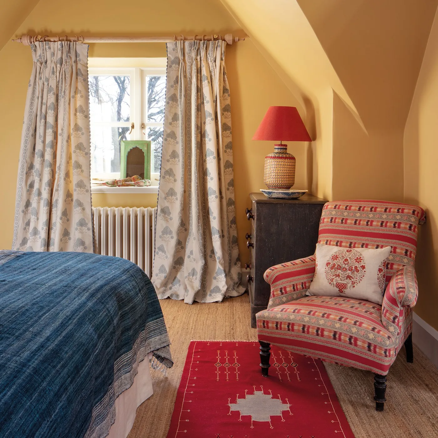 Warm yellow bedroom with a blue bed cover, patterned curtains, red-striped armchair and a small dark chest.