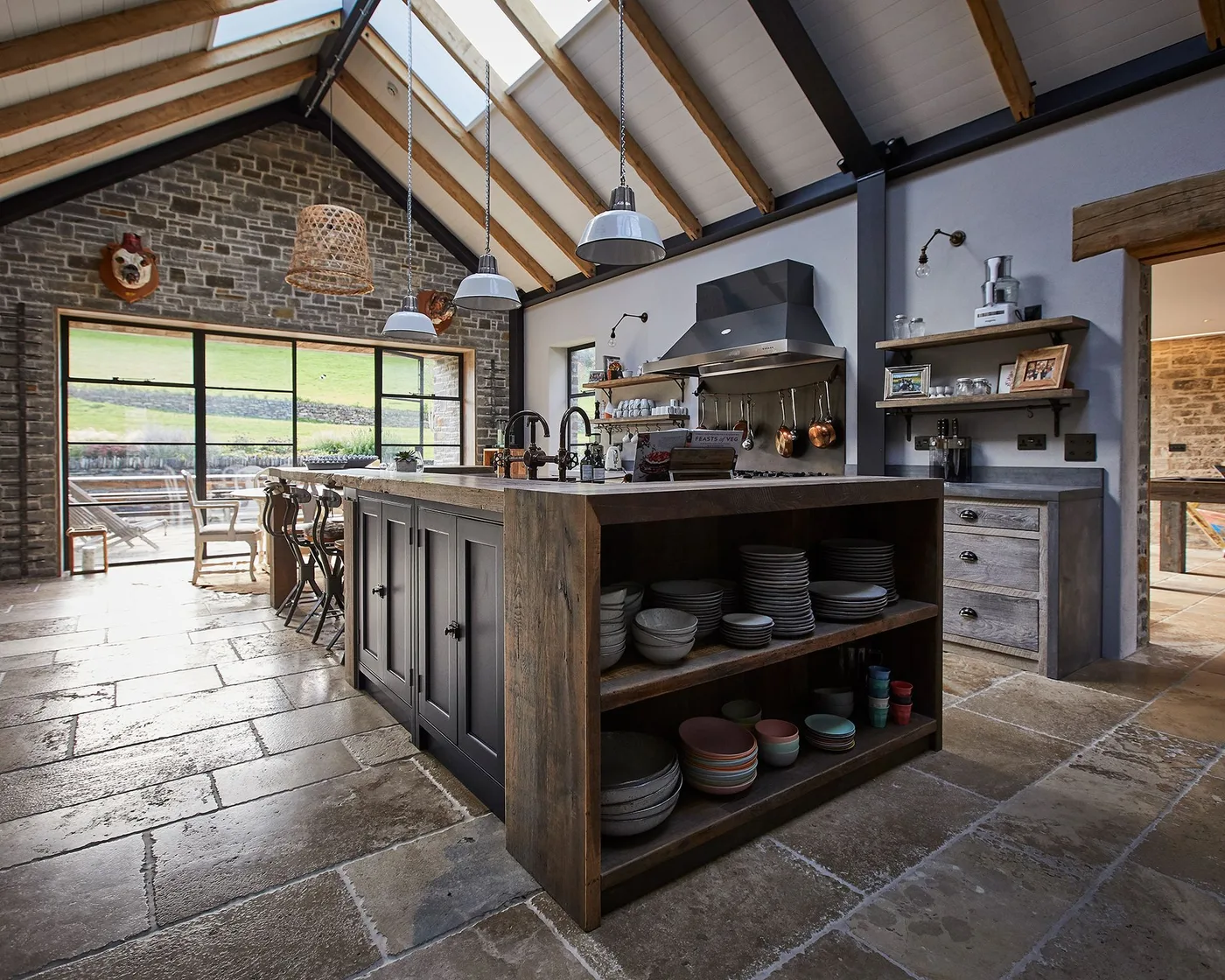 Kitchen with dark wood island, open dish shelves, gray wall cabinets, brick end wall, high timber-and-metal roof, hanging lights, and large glazed doors.