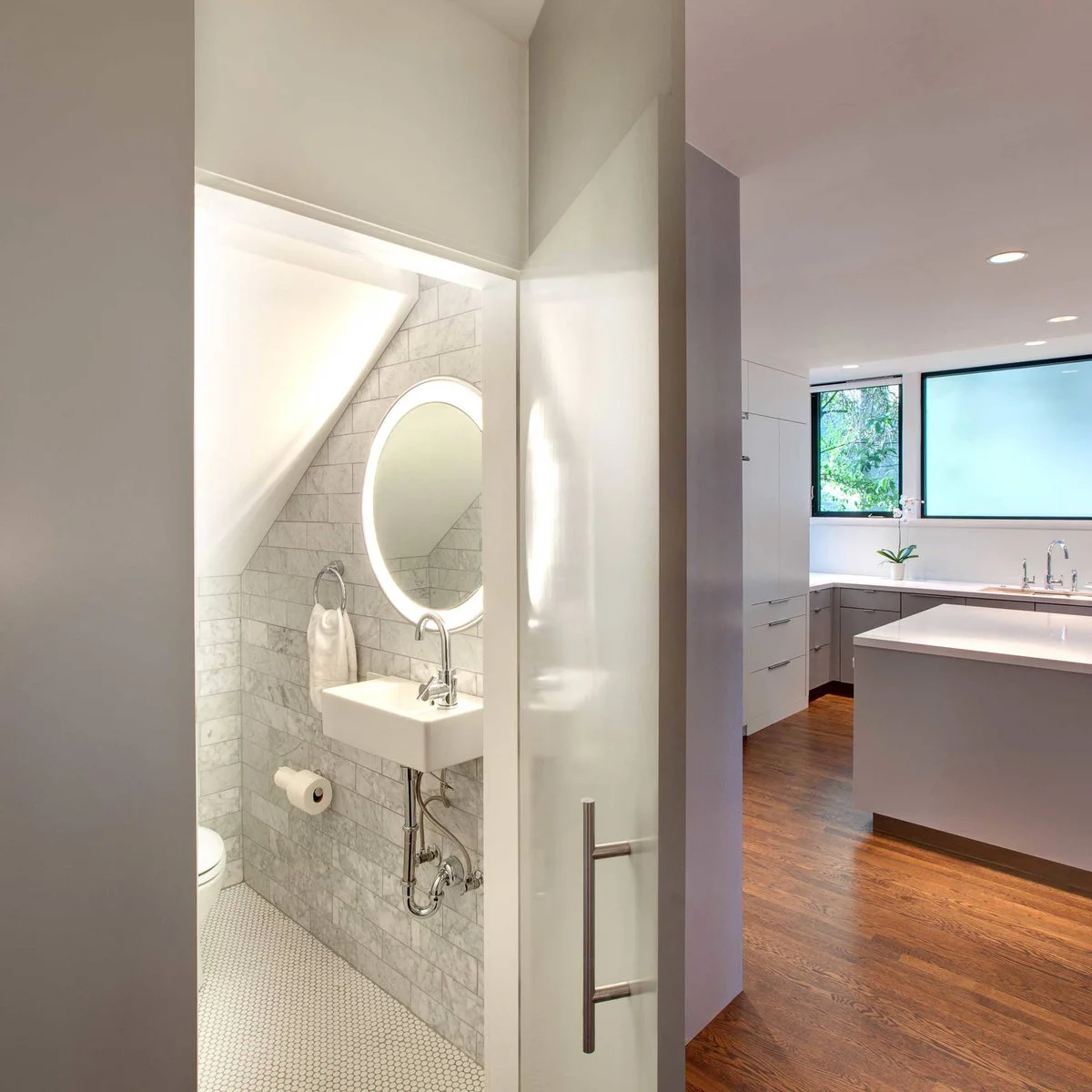 Illuminated round mirror above a small wall-mounted sink beneath a sloping ceiling