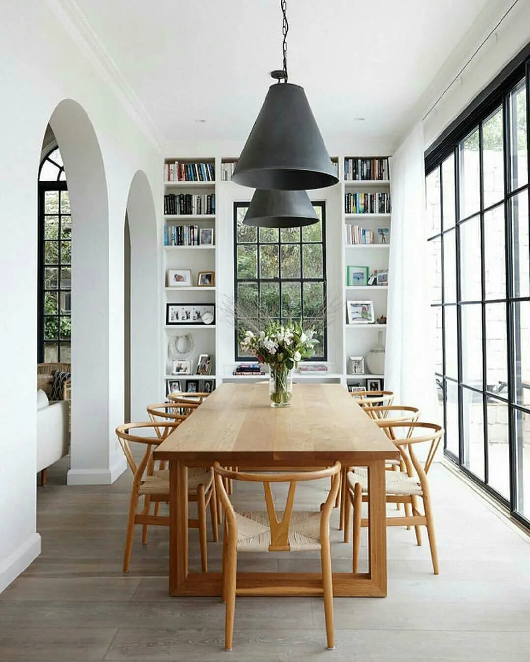 Dining room with rectangular wooden table, curved wooden chairs, black pendants, tall black-framed glazing and built-in bookshelves.