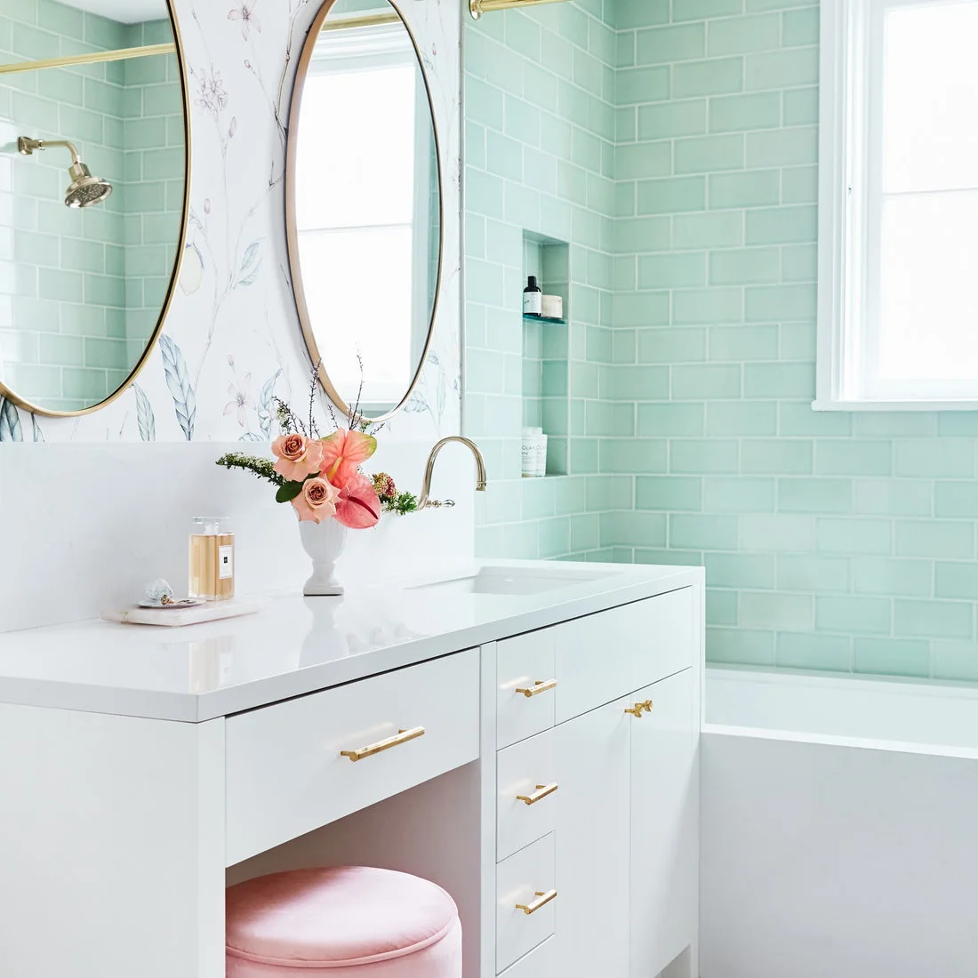 A pink upholstered stool sits beneath a white vanity beside pale green shower tiles and floral wallpaper.