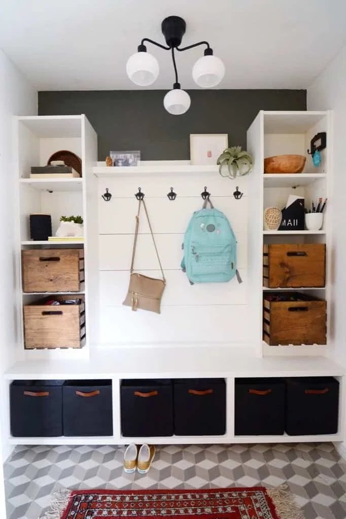 White entryway storage with two open side units, wood boxes, black lower baskets and a central row of hooks.