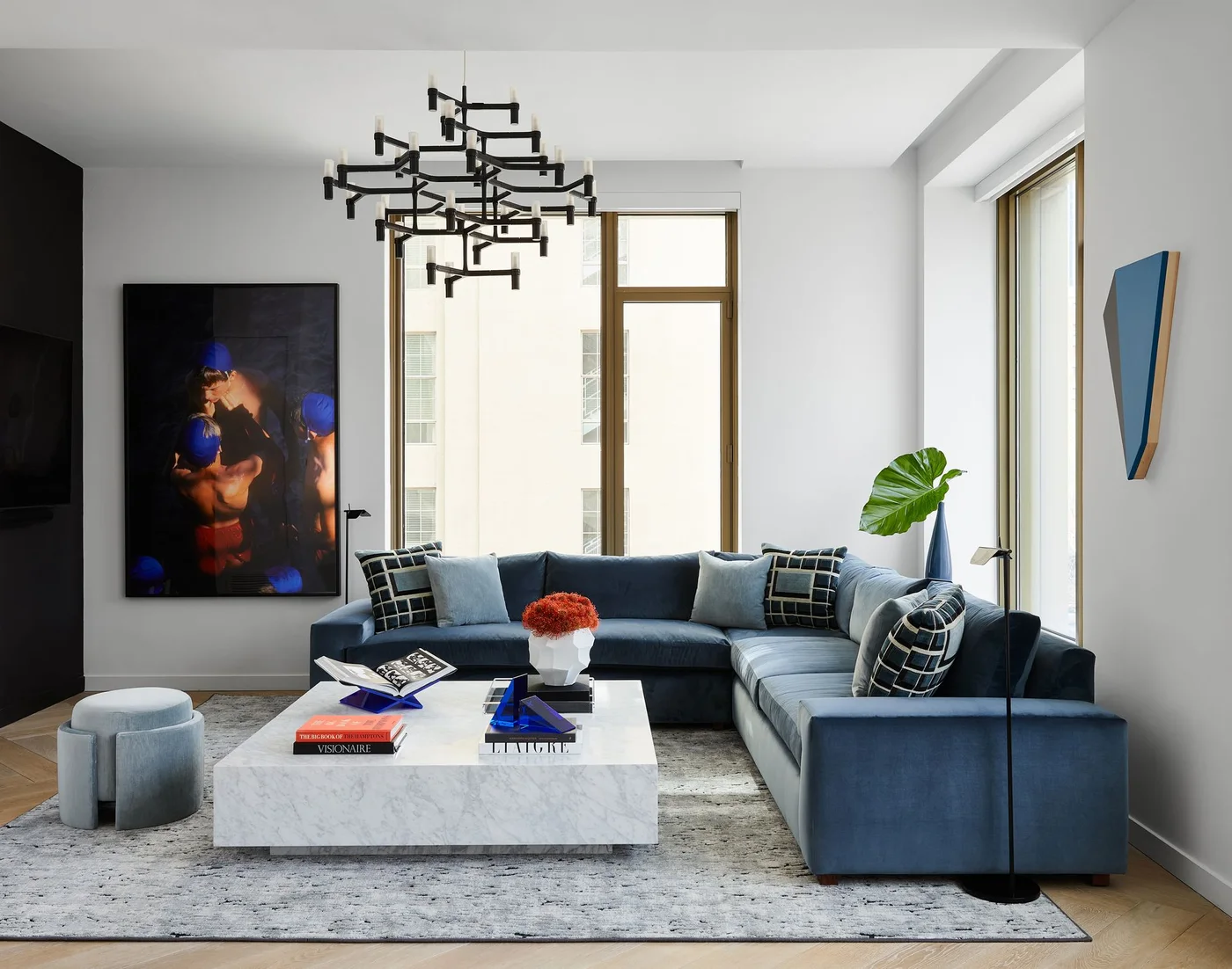 A blue sectional sofa surrounds a pale low block-shaped coffee table beneath a dark branching light beside tall windows and large artwork