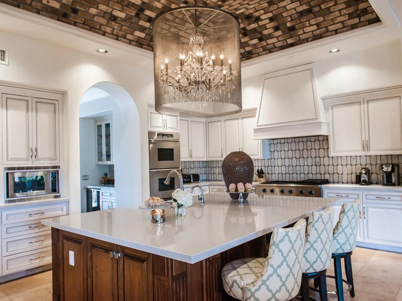 White kitchen cabinets surround a timber island below a vaulted tiled ceiling and a chandelier enclosed by a broad shade.