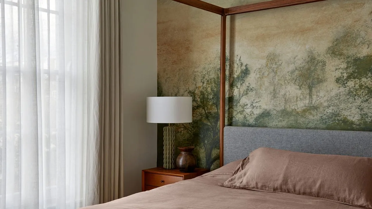 A soft landscape mural rises behind a pale headboard beside light curtains.