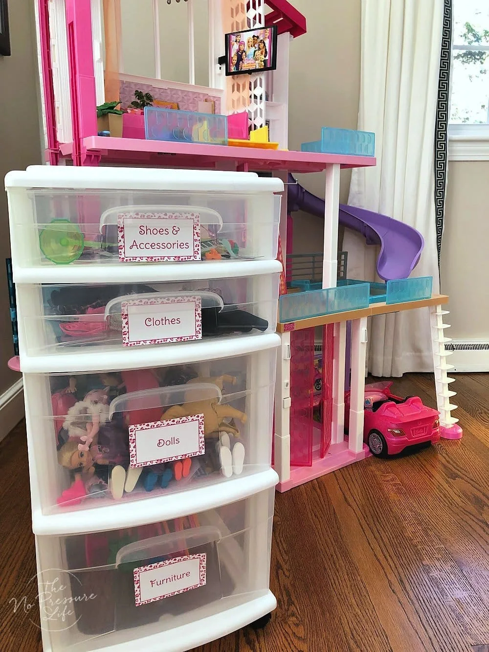 White-framed clear drawer tower labeled Shoes and Accessories, Clothes, Dolls and Furniture beside a large pink dollhouse.