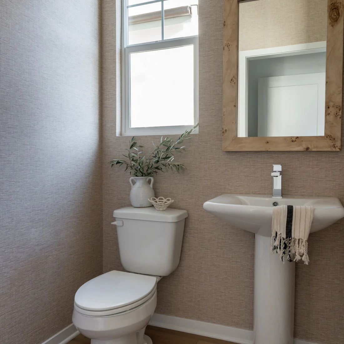 A white pedestal basin and toilet sit against finely textured beige walls beneath a wood-framed mirror and a small high window.