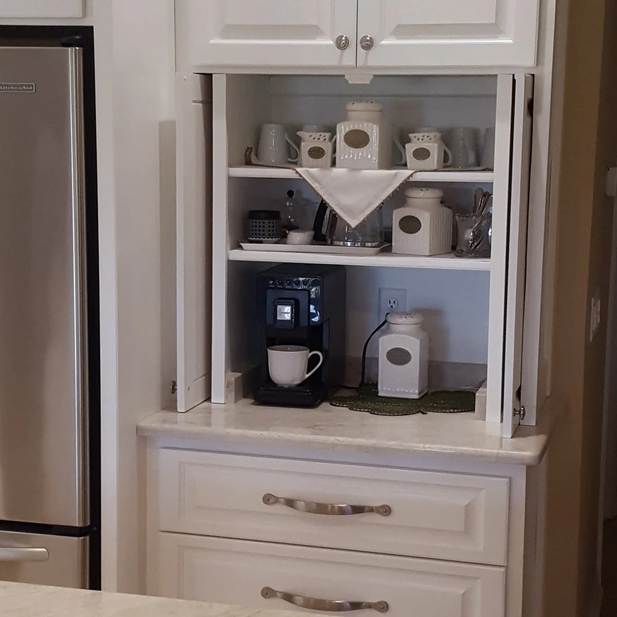 White countertop coffee cupboard with doors folded back beside a refrigerator