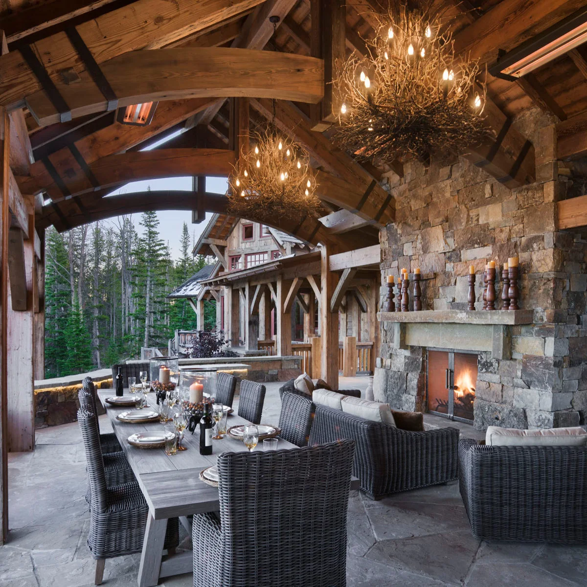 Two branch-like chandeliers hang above a long dining table under a vaulted timber patio roof beside lounge seating.