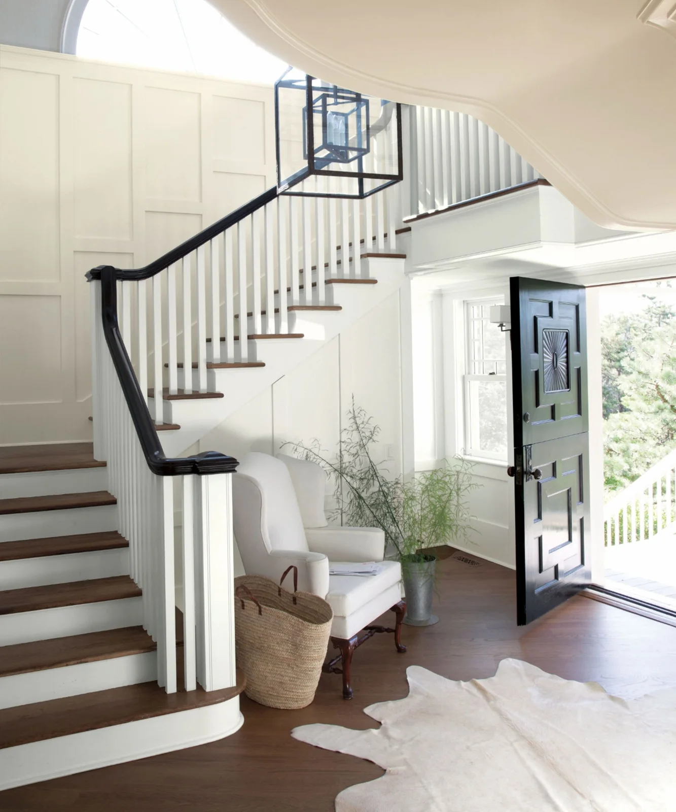 A pale paneled entrance has a dark front door and black stair handrail above white balusters and wood steps beside a light chair