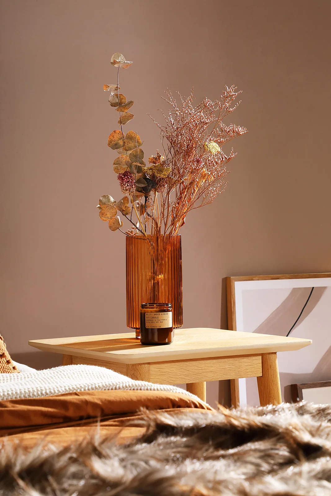 An amber ribbed vase holds a light arrangement of dried flowers and round leaves on a bedside table beside pale bedding and a brown wall.