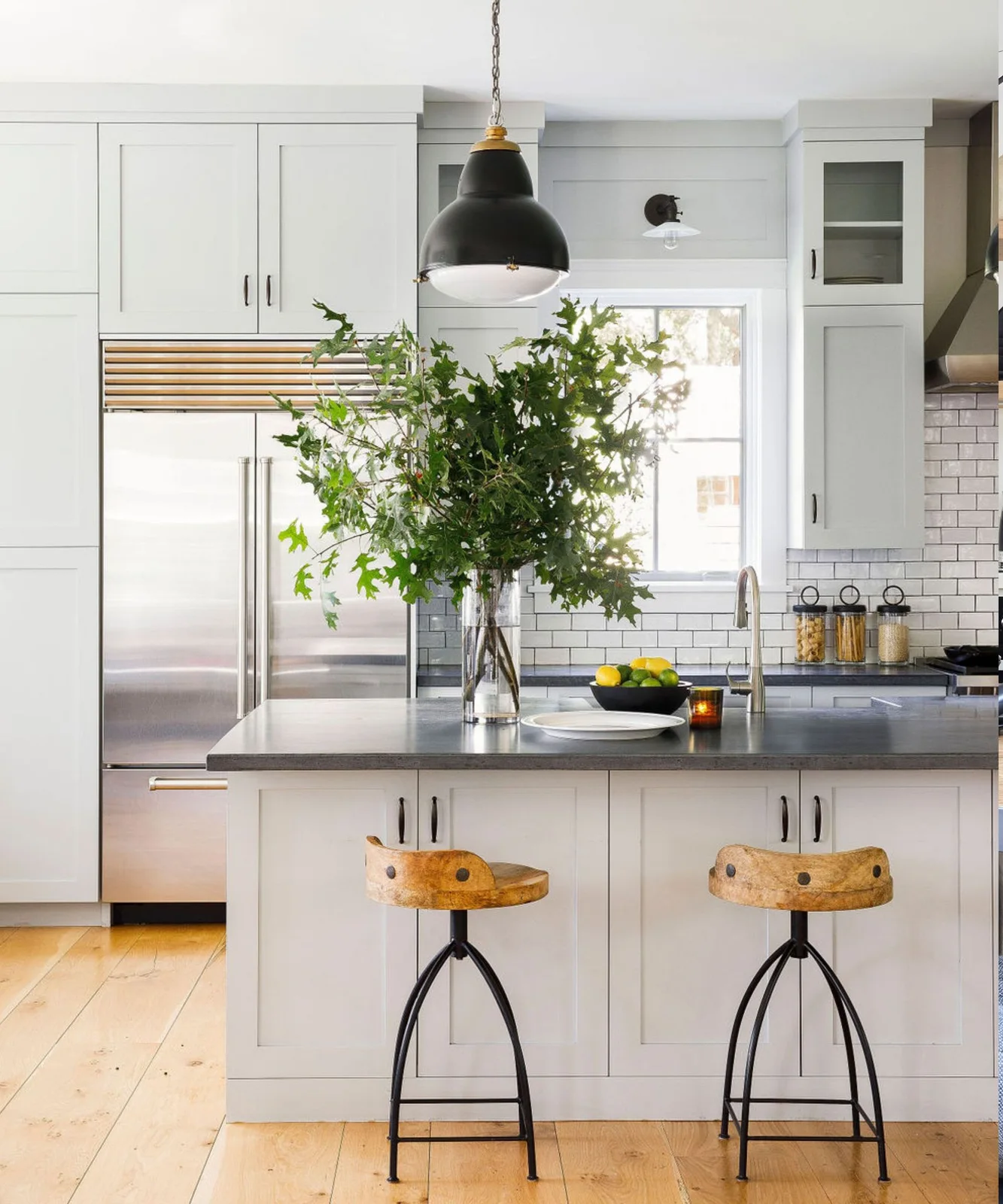 A white framed kitchen has a dark island counter, black cabinet handles, wood-and-metal stools, a black dome pendant and a metal refrigerator.