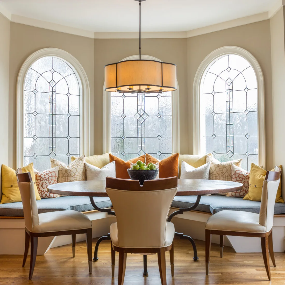Oval dining table with pale chairs in front of a cushion-filled curved window bench and arched windows