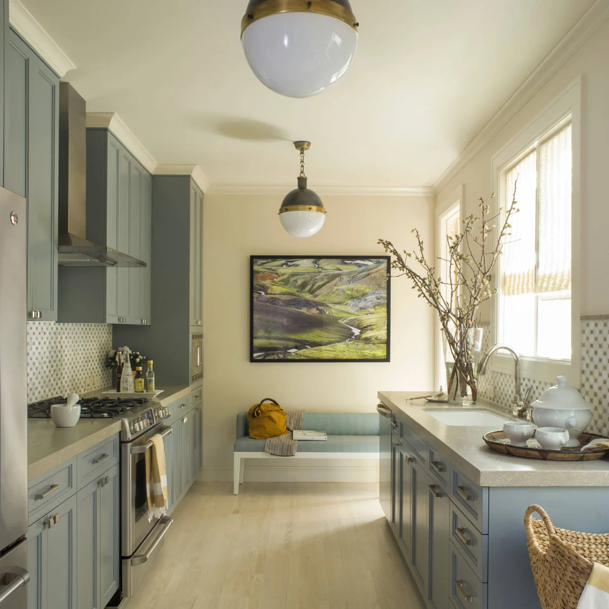 Gray cabinets line a narrow kitchen with cream walls, pale counters, a small mosaic backsplash and a pale upholstered bench.