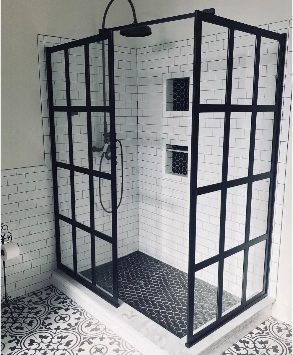 Two black grid shower screens frame an open corner above a white curb, with black hexagonal flooring and white subway walls.