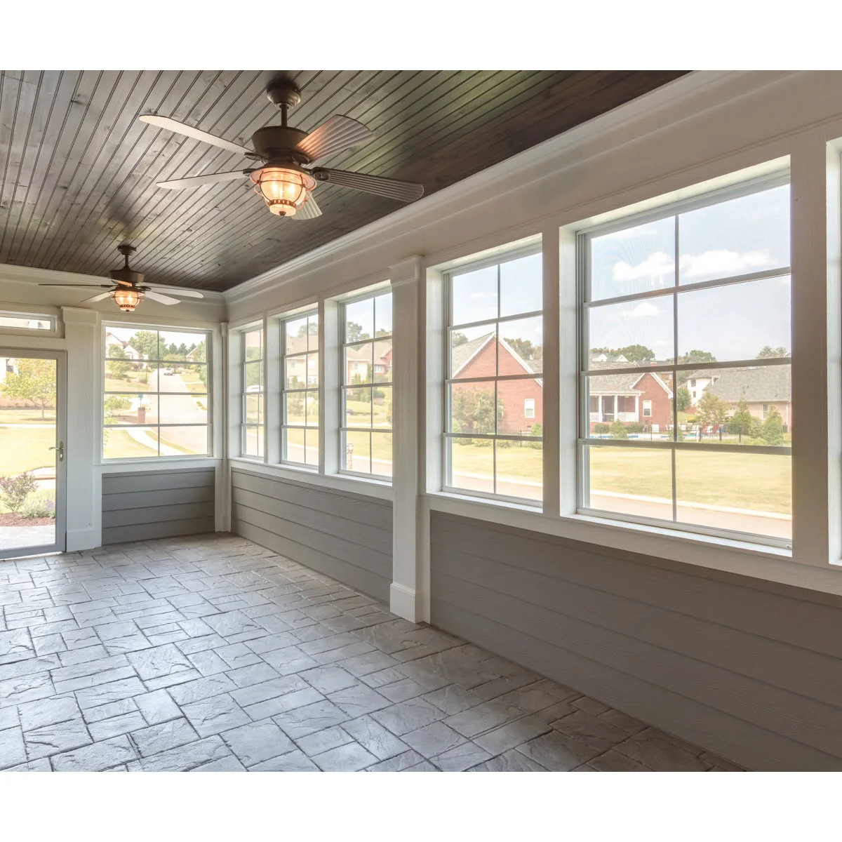 An unfurnished enclosed porch with pale patterned flooring, white-framed windows, gray lower walls and a dark wood ceiling.