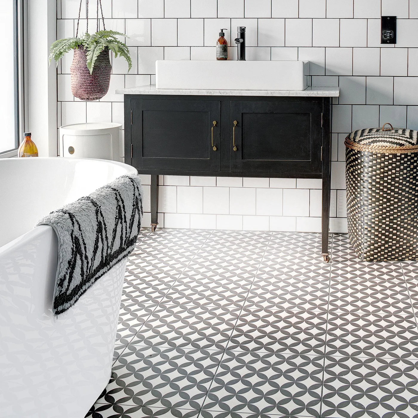 A black vanity on small warm-metal legs supports a white rectangular vessel basin, beside white tiled walls, a patterned floor and a woven hamper.