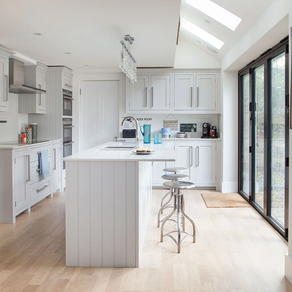 White framed cabinets surround a narrow island with vertically paneled ends and metal stools beside broad garden doors.