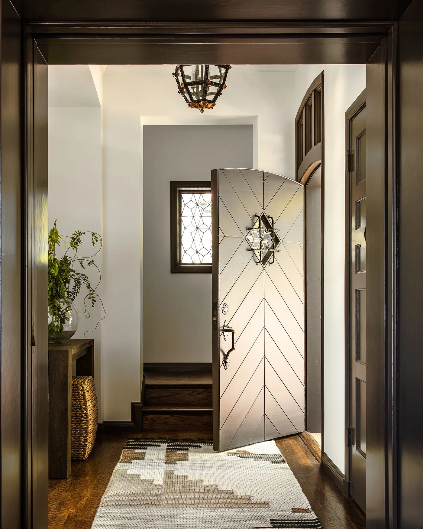 Neutral geometric entrance rug beside a diagonally detailed door, dark trim, narrow console, small window and hanging lantern