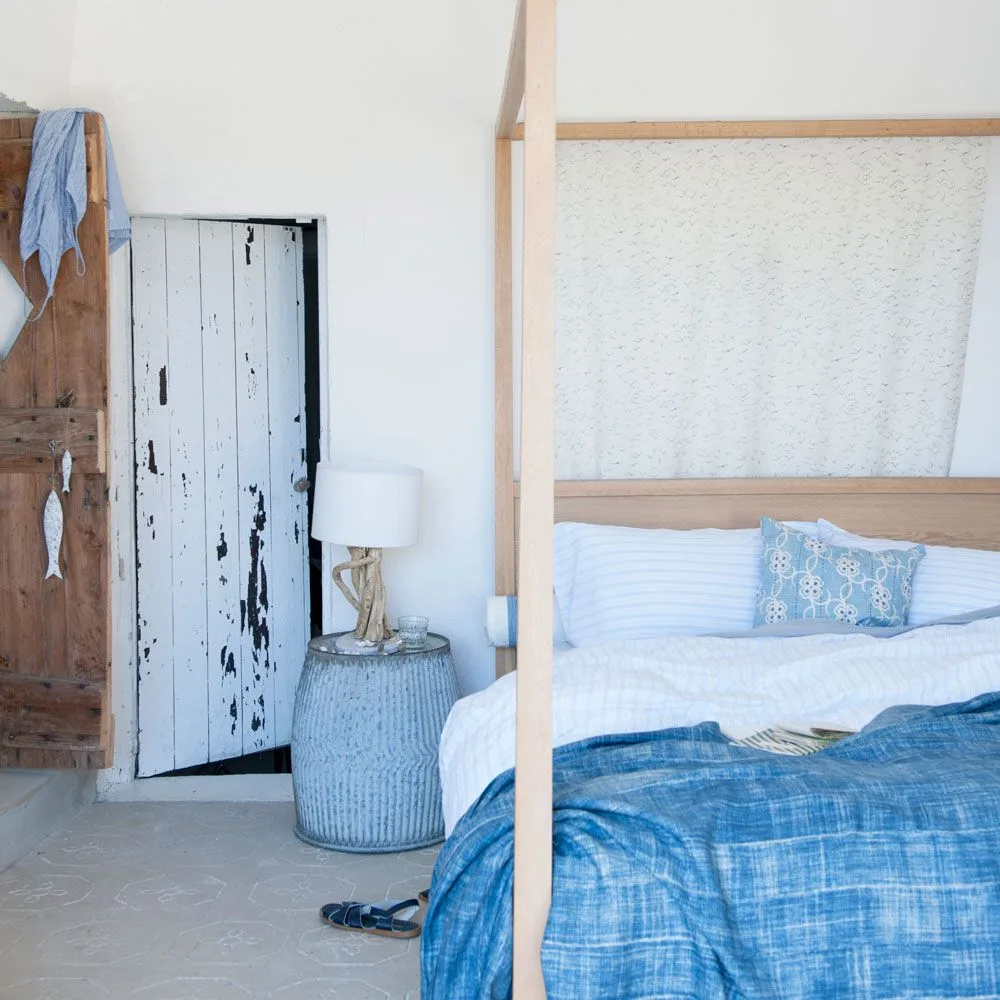 Pale bedroom with a simple wooden post bed, white-and-blue bedding, blue round bedside table and a distressed-looking painted door.