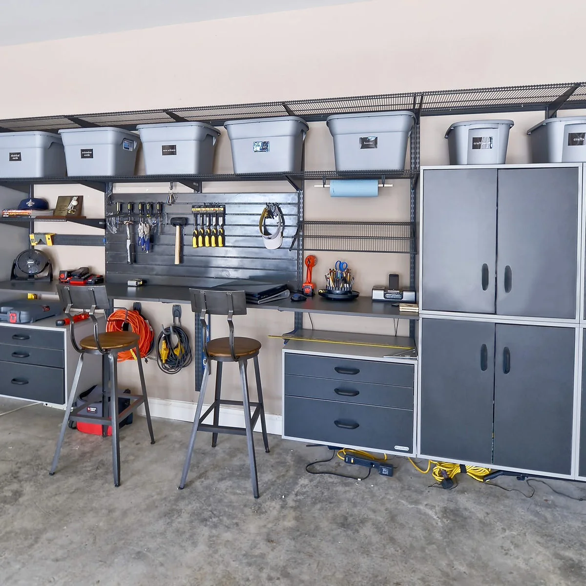 Long garage workbench with gray cabinets, two stools, small tools on a wall panel and labeled storage tubs above.