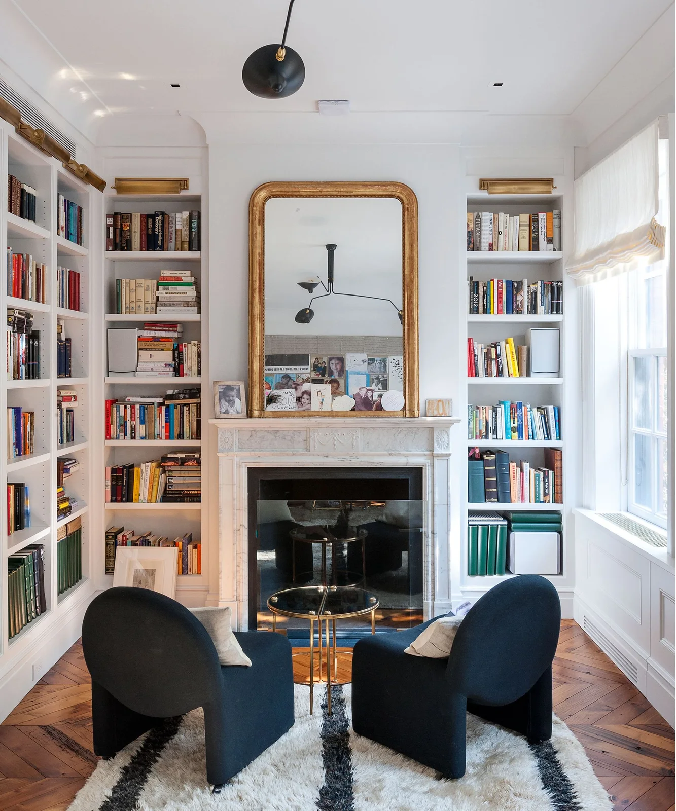 White bookcases frame a fireplace and gold-toned mirror beside two dark blue chairs on a patterned rug