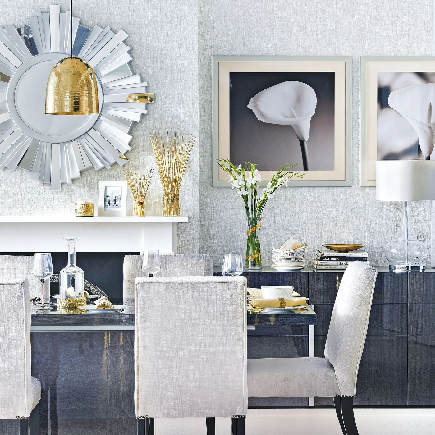 A dark reflective sideboard sits behind pale dining chairs beneath framed floral pictures and a clear lamp beside a pale wall