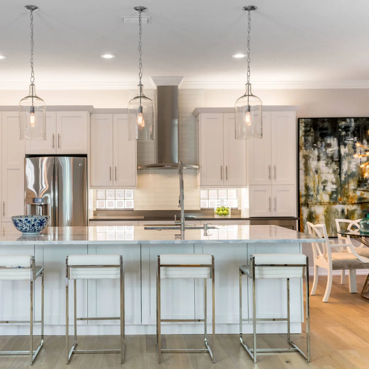 Three narrow clear glass pendant shades hang above a pale island with upholstered stools and silvery frames.