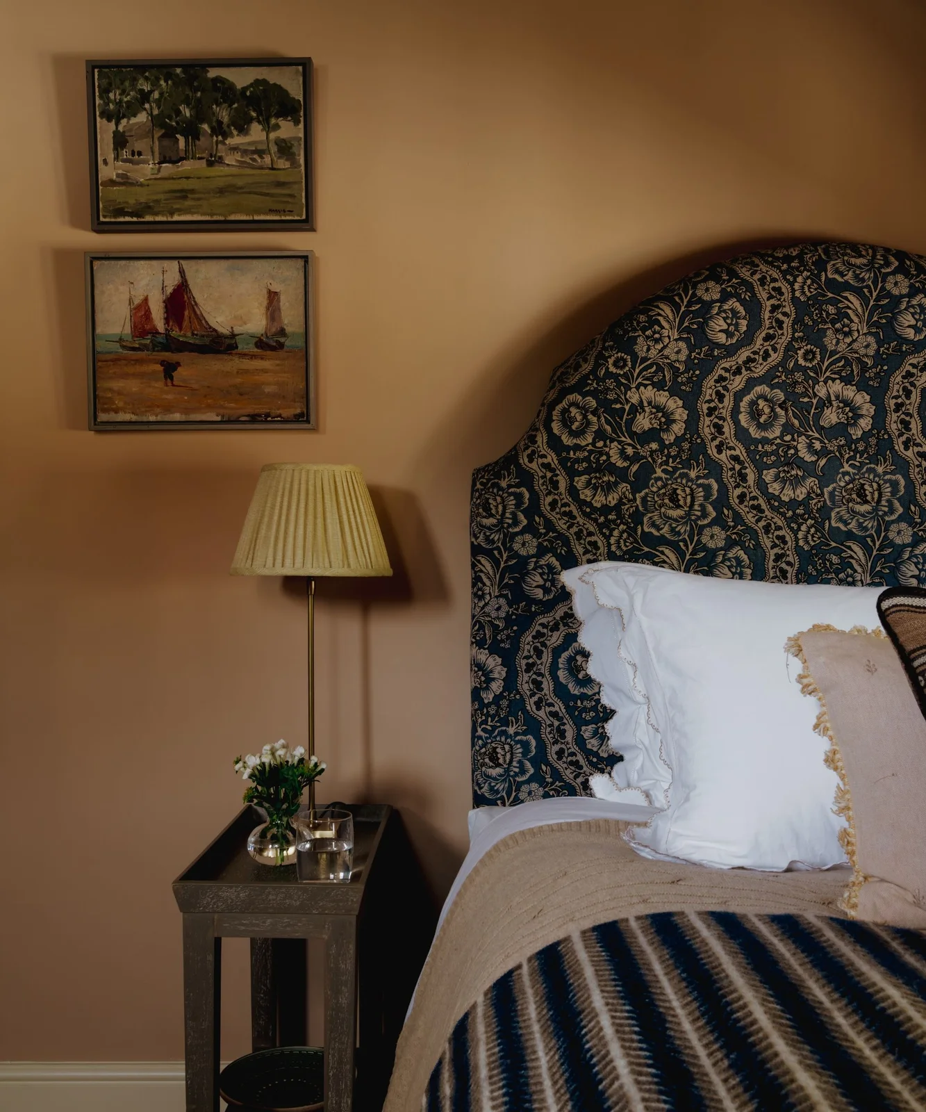 Muted apricot bedroom wall behind a blue floral headboard and pale bedding with a striped throw.