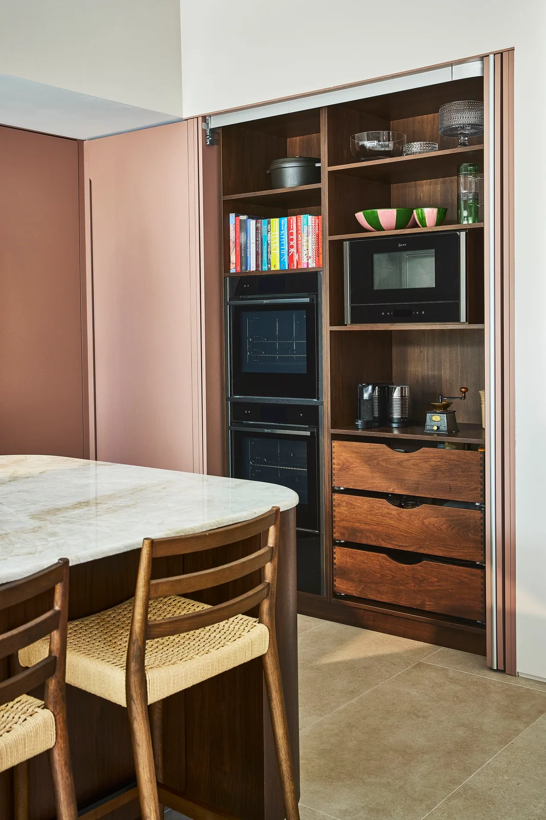 Open pocket doors reveal walnut shelves, drawers and black appliances within a dusty pink tall cabinet wall.