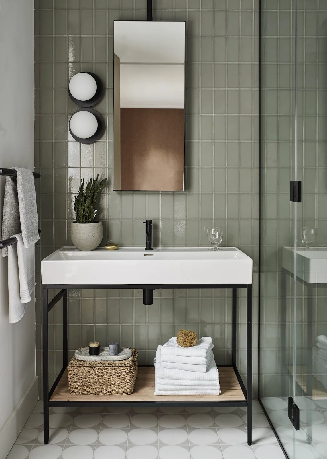 Gray-sage vertical tiles frame a white console basin on a black stand with a wood shelf, narrow mirror and twin globe light.