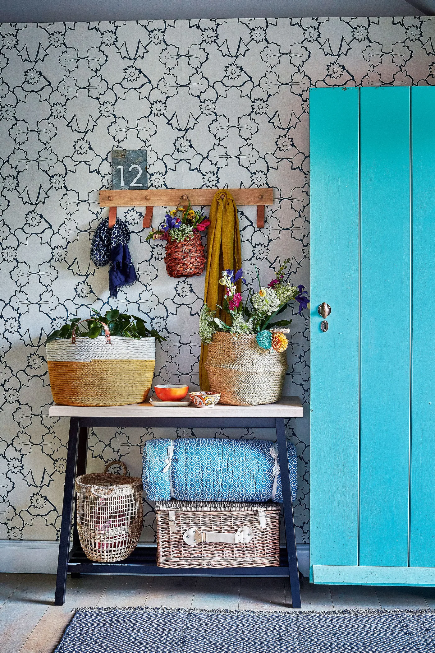 Entrance with a bright turquoise plank door, black floral wall pattern, small dark console, woven baskets and a wooden hook rail.