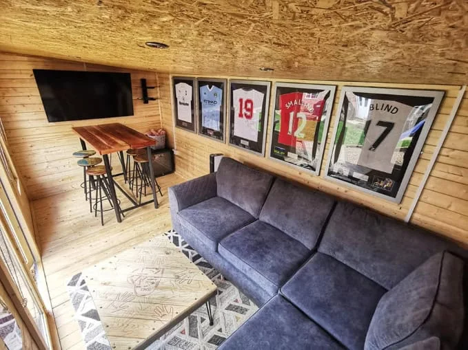 Timber garden room with a grey sofa, a high table, stools and framed sports shirts
