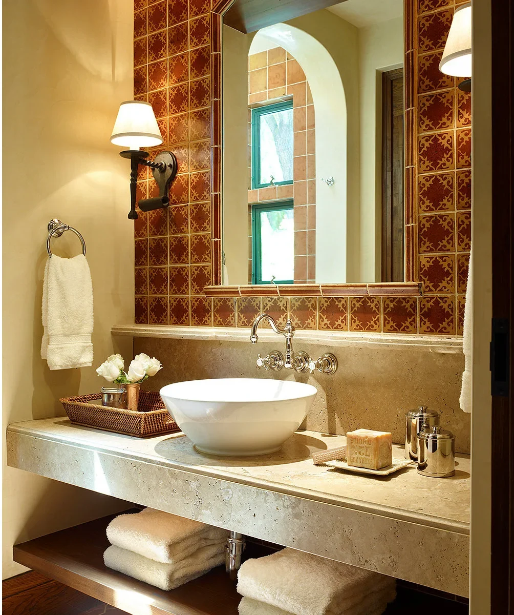 A white vessel bowl sits on a pale counter beneath wall-mounted chrome fittings and a mirror framed by warm patterned tiles.