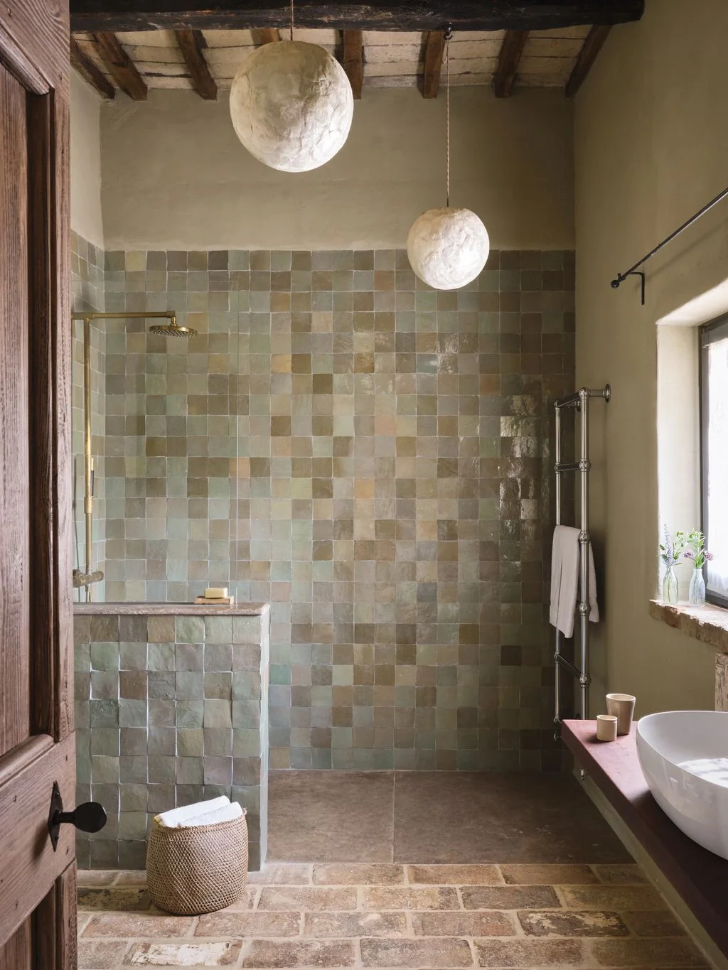 A low tiled wall shelters an open shower with muted green and beige square tiles, exposed beams and pale rounded pendant lights.