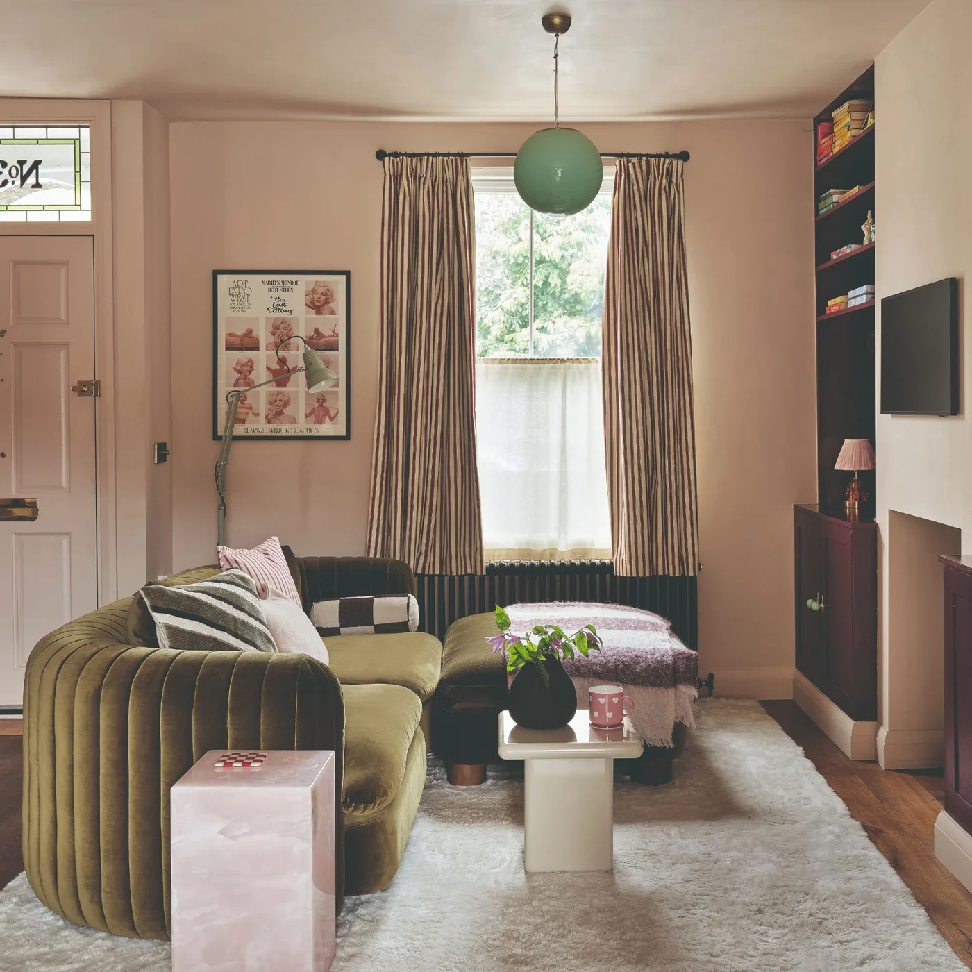 An olive curved sofa and small pale tables sit on a light rug in a narrow pink room with dark alcove storage.