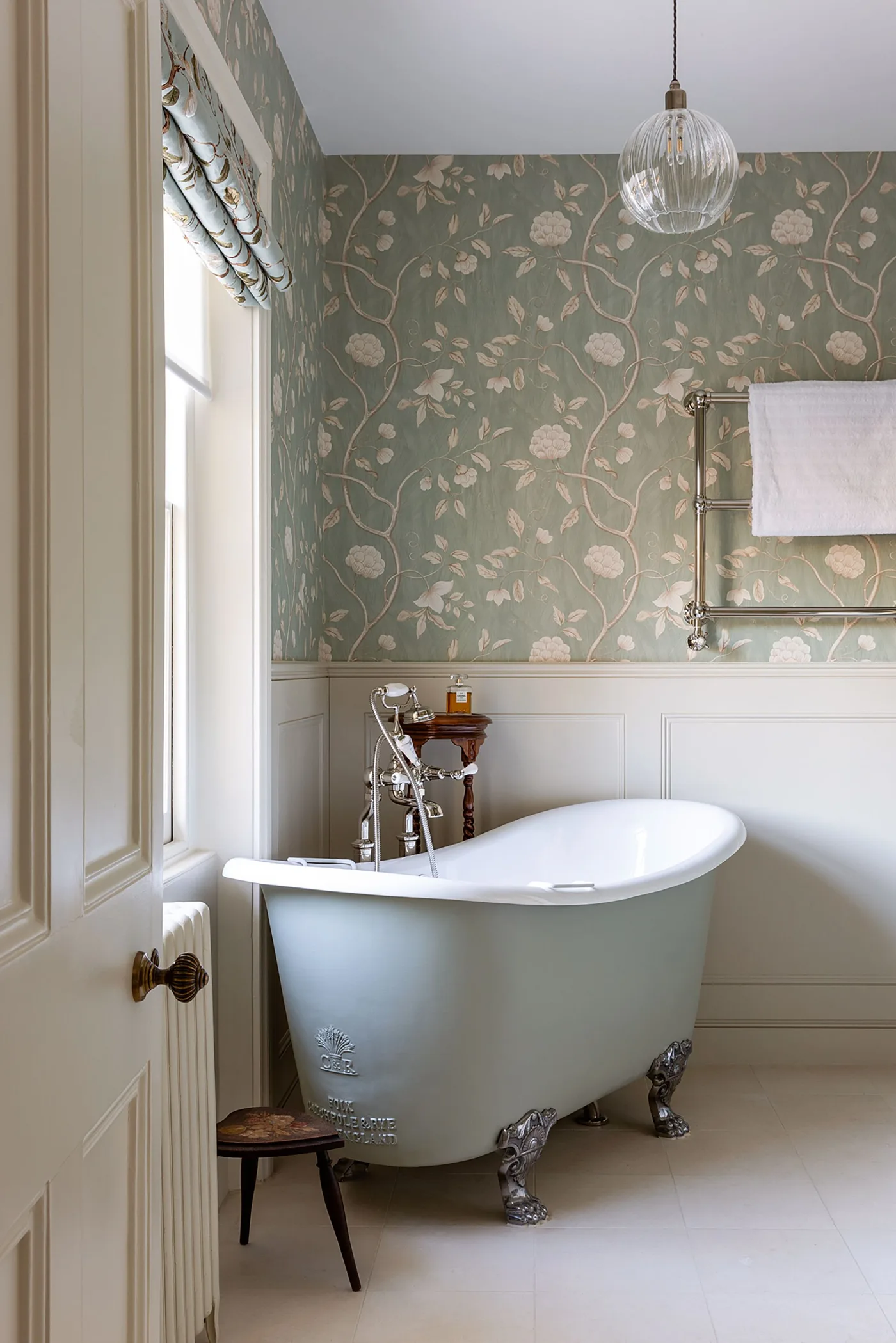 Small bathroom with pale green floral wallpaper, white lower paneling, a pale claw-foot bath and a fabric window blind.