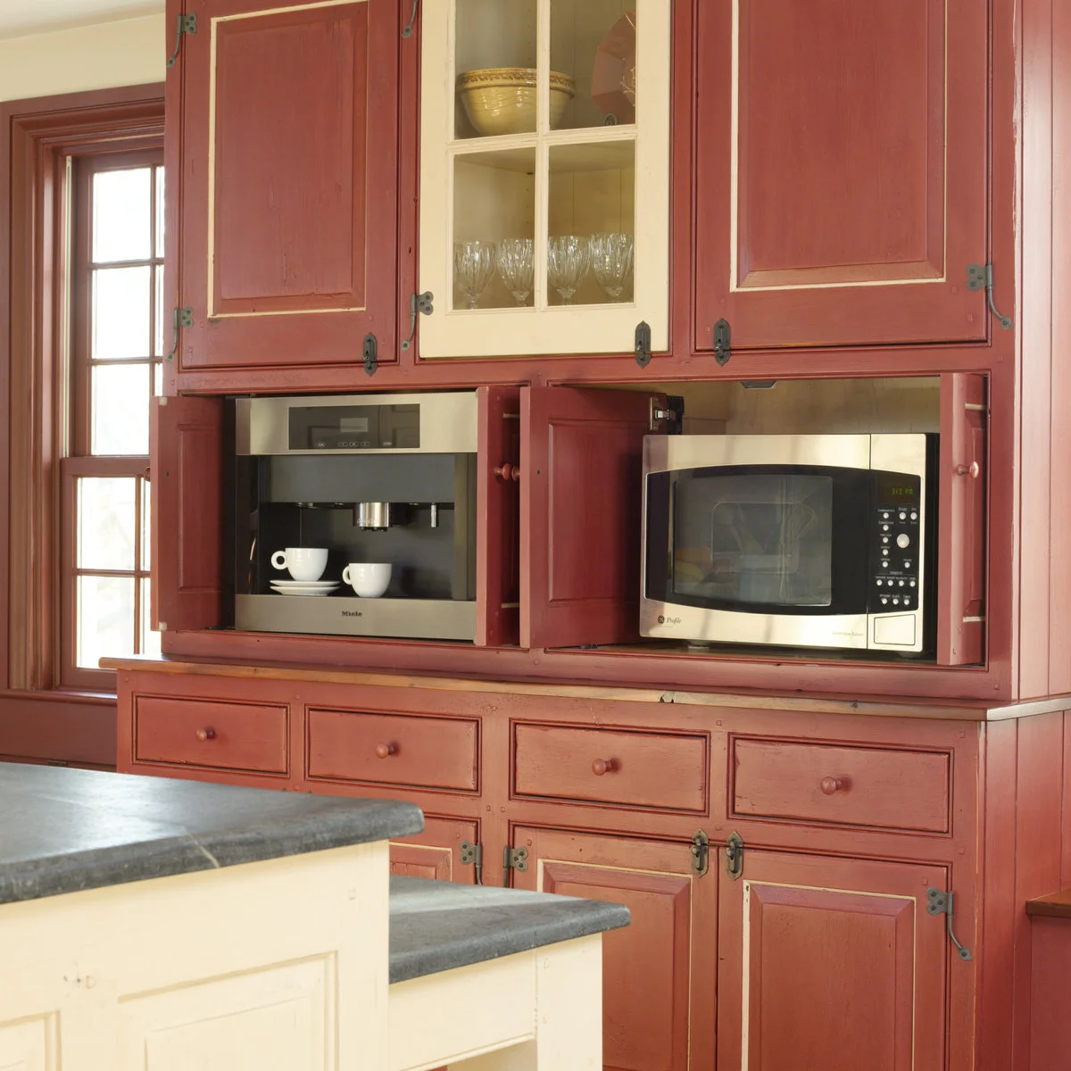 Muted red cabinets with dark decorative hinges surround a cream glass-front door and appliances beside cream islands with gray tops.