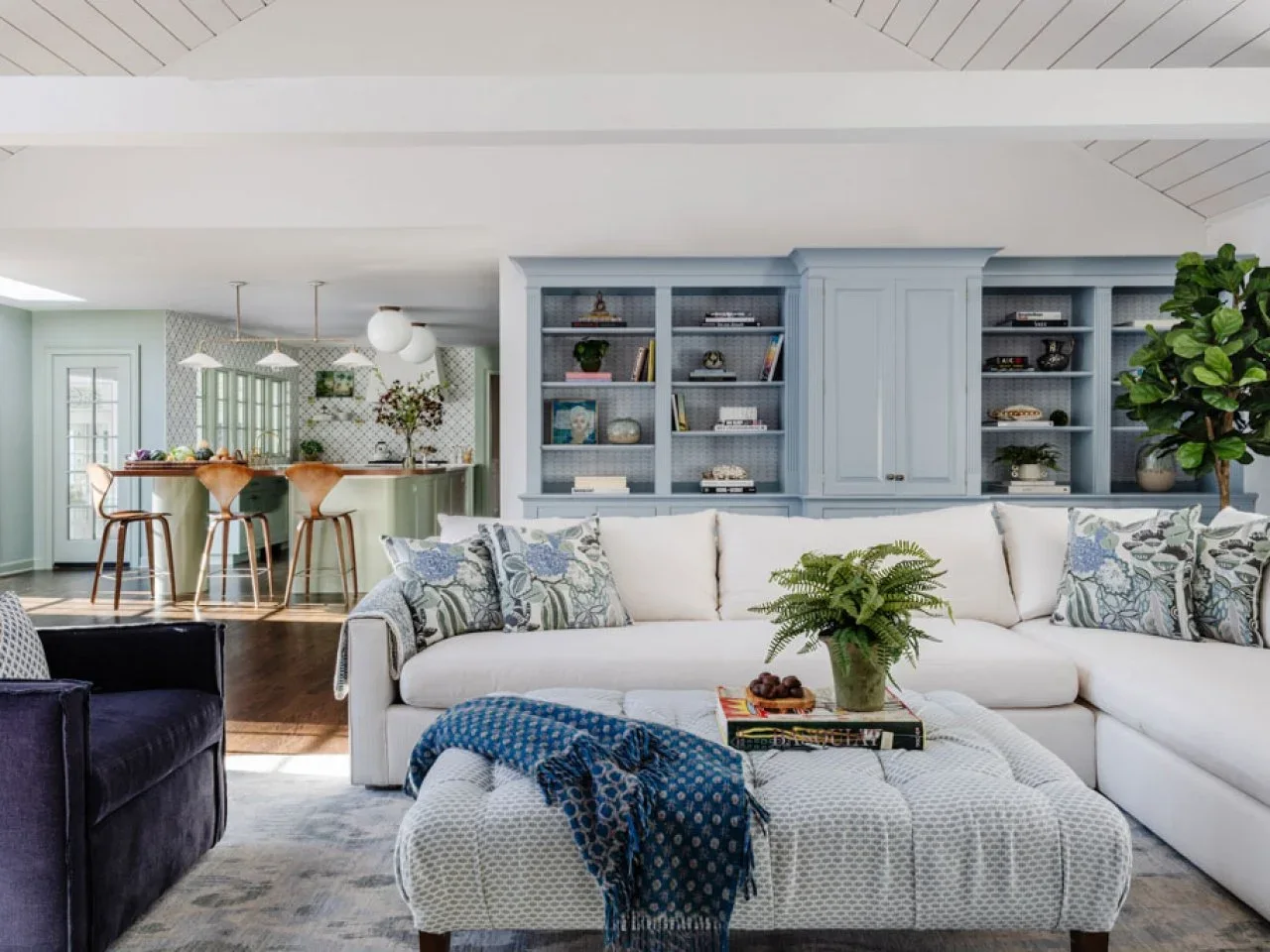 A white sectional sits before pale blue built-in shelves with an open kitchen and timber breakfast counter beyond.