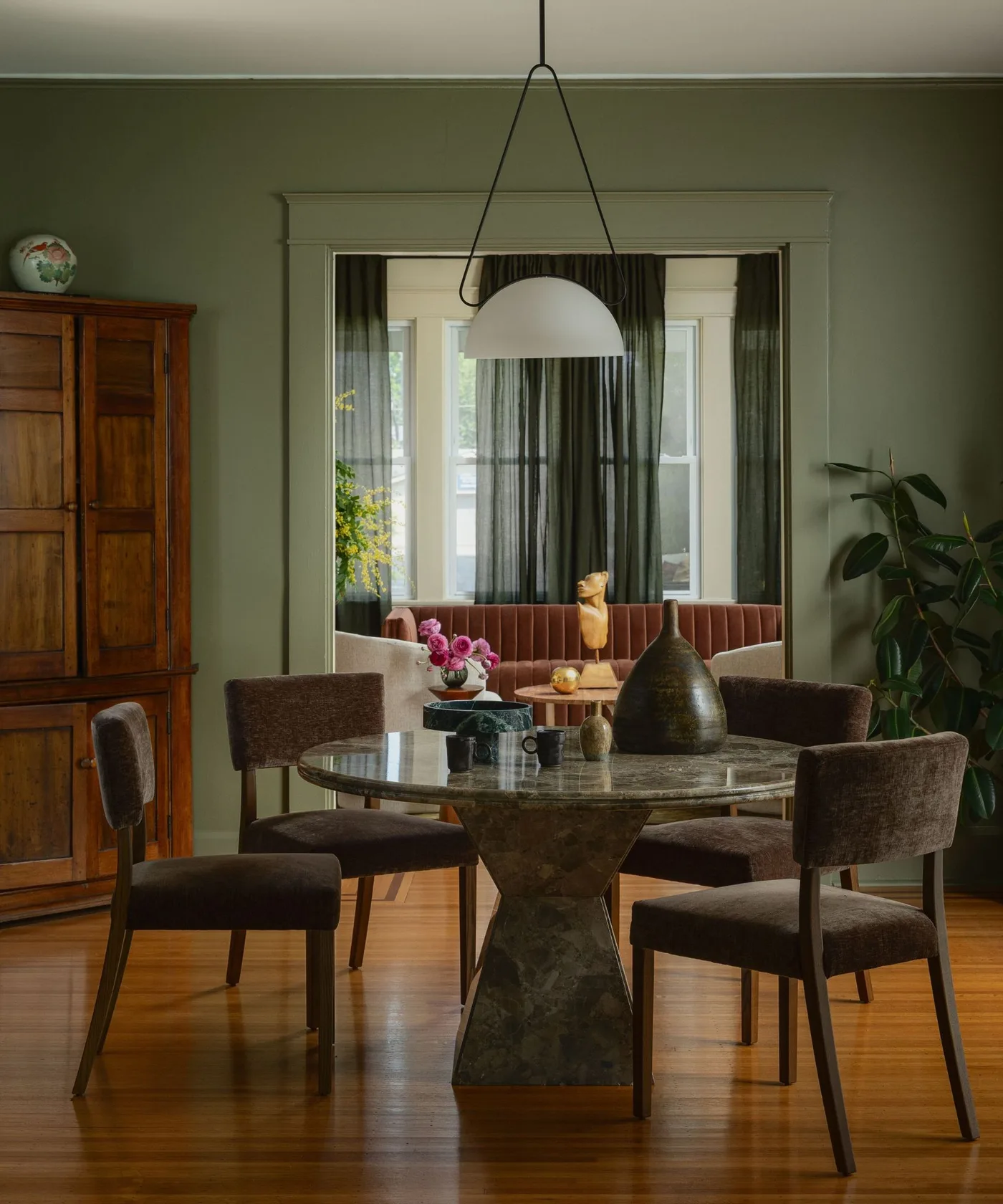 Green dining room with a tall wooden cabinet, glossy round pedestal table, brown upholstered chairs and a large mirror.