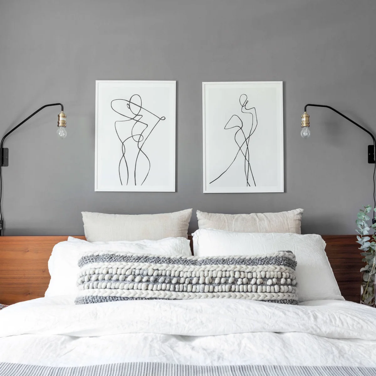 Two white-framed line drawings above a wooden headboard on a gray wall with curved black bedside lights.