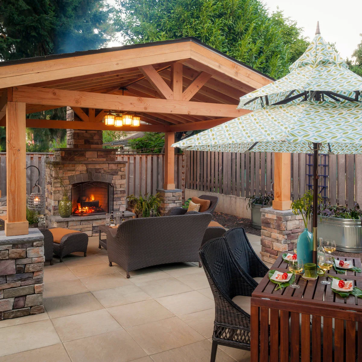 A wood pavilion on stone-clad piers shelters seating by a fireplace, with a dining table under a patterned umbrella outside.