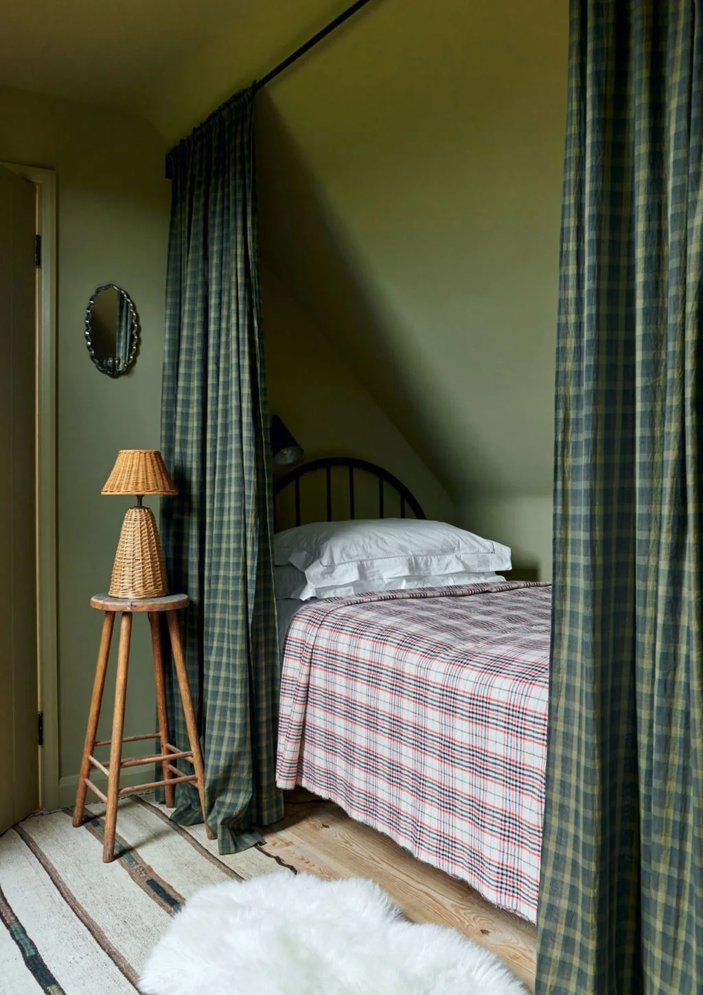 Small green bedroom nook with checked curtains, checked bed covering and a woven lamp on a stool