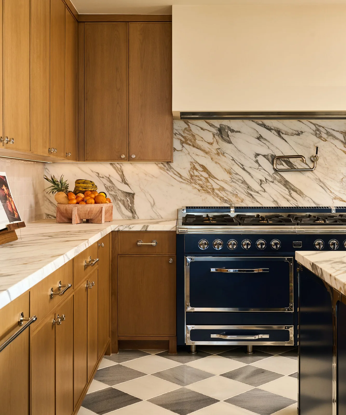 Honey-toned oak cabinets frame a navy range beneath a pale hood, with veined counters and a gray-and-white checked floor.