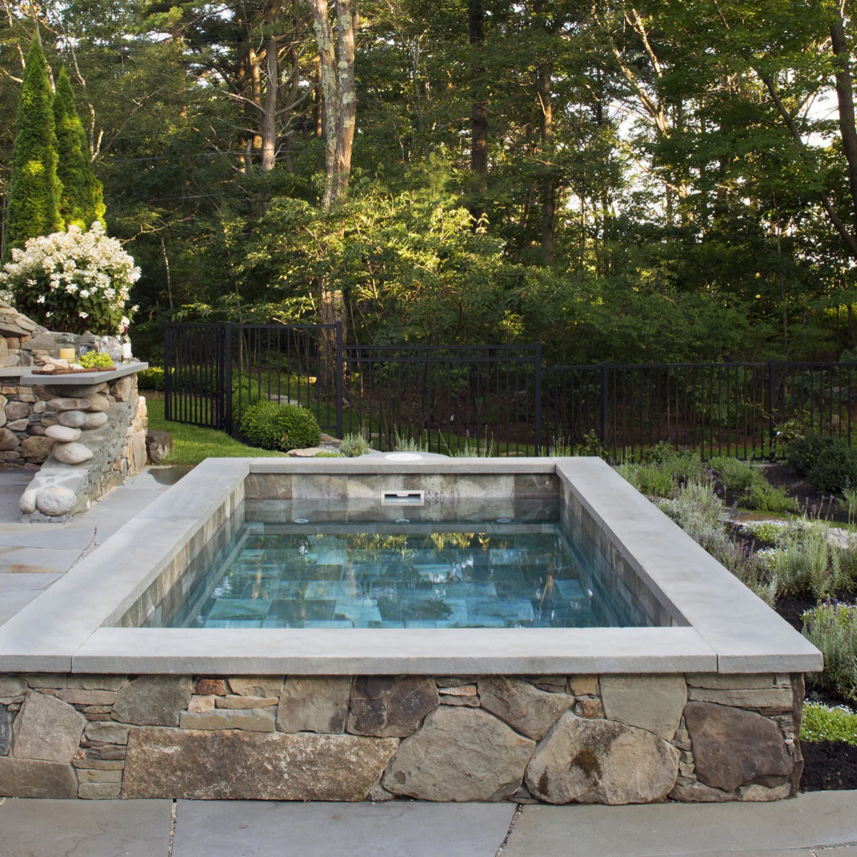 A small raised stone-faced pool sits beside a matching outdoor counter, gray paving, low planting and a dark metal garden fence.