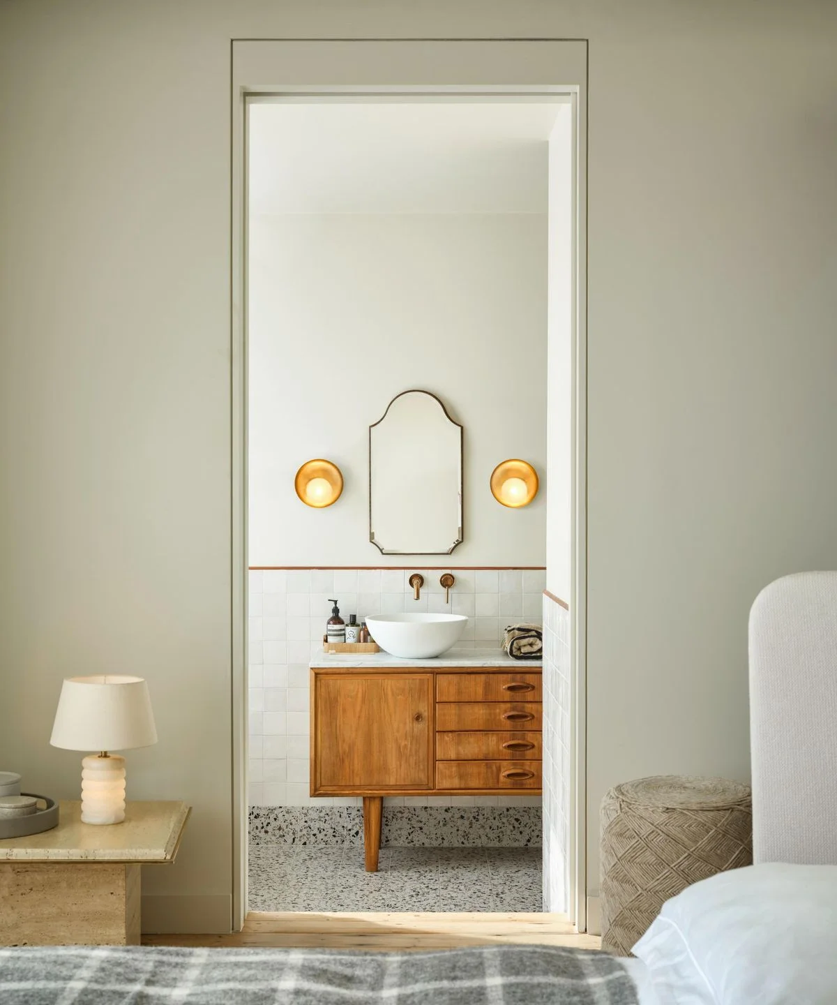 A bedroom doorway opens onto a wood drawer vanity with a white vessel bowl, shaped warm-metal mirror, dome wall lights and a speckled floor.